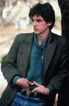 a man in a green shirt and jacket leaning against a tree with his hands in his pockets