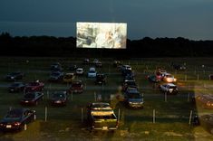 cars parked in a lot at night with a large screen on the wall behind them