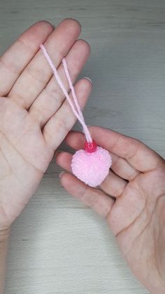 two hands holding a small pink pom - pom ornament on a string