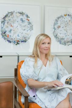 a woman sitting in a chair reading a book with paintings on the wall behind her