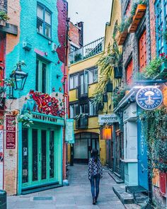 a woman walking down an alley way in front of colorful buildings with plants growing on the windows