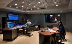 two men sitting at desks in an office setting with monitors and sound equipment on the walls