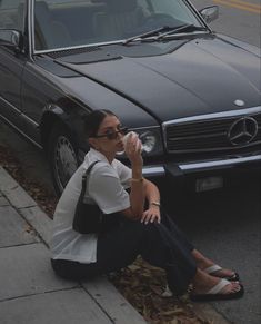 a woman is sitting on the curb drinking from a cup in front of her car