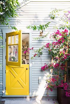 a yellow door is open in front of a gray house with pink flowers on the side