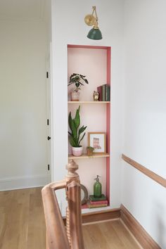 a pink shelf with books and plants on it next to a wooden stair case in a white walled room