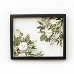 a black frame with white flowers and green leaves on the inside is hanging on a wall