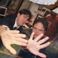 two men sitting at a table with their hands in front of them and one holding out his hand to the camera