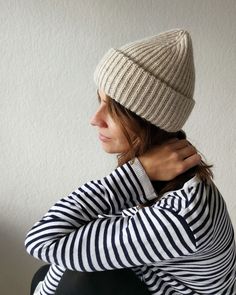 a woman wearing a white and black striped hat with her hands on her chest, sitting against a wall