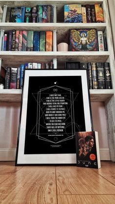 a book shelf filled with books next to a white framed poster on top of a wooden floor