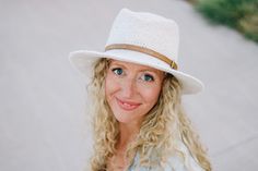 a woman with blonde hair wearing a white hat and smiling while standing on the sidewalk