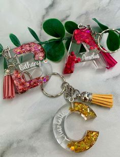 three personalized keychains with tassels and charms on top of a marble surface