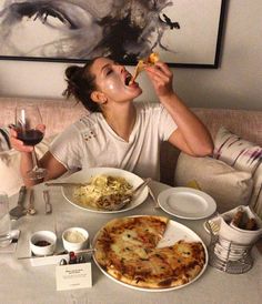 a woman sitting at a table eating pizza and drinking from a wine glass in front of her