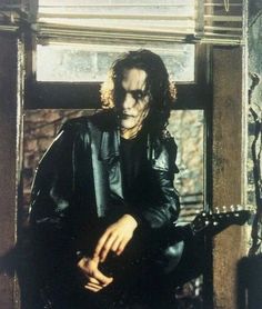 a man with long hair and makeup sitting in front of a window holding a guitar