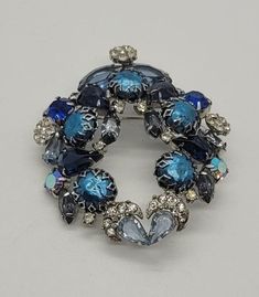 Feeling blue? This vintage brooch is for you! There are three different shades of blue rhinestone crystals in this unusual design, which are accented by the five, prong-set faux Baroque pearls. I believe this is an unsigned piece of Juliana costume jewelry, produced by DeLizza & Elster, which was founded in 1947. I am compelled to see this as a designer piece, given its unique wreath design and multitude of cuts and colors. There are separate clusters of clear crystals on three sides and lovely caps of crystals atop the inverted teardrops at the base. According to Collectors Weekly, Juliana manufactured everything from buttons and buckles to pins and pendants for such venerable costume jewelers as Hattie Carnegie, Hobe, Kenneth J. Lane, and Weiss starting in the 1940s. In 1967, chief desig Unique Blue Brooches For Formal Occasions, Unique Blue Brooch For Party, Unique Blue Brooches For Party, Blue Vintage Brooch For Party, Costume Jewelry Blue Brooches With Rhinestones, Antique Blue Jewelry With Brooch, Vintage Blue Jewelry Brooch, Vintage Blue Brooch, Blue Crystal Jewelry Brooch