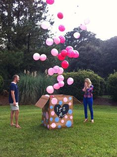 a man and woman standing in the grass near a cardboard box with balloons floating from it