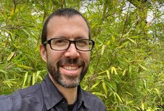 a man wearing glasses standing in front of some trees and bushes smiling at the camera