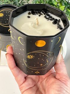 a hand holding a black cup filled with white liquid and stars on the side, in front of a potted plant