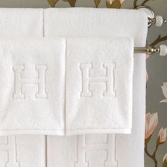 two white towels with the letter h on them sitting next to each other in front of a floral wallpaper