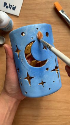 a hand holding a paintbrush in front of a cup with the moon and stars painted on it