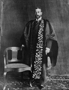 an old photo of a man wearing a robe and standing in front of a chair