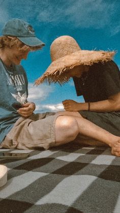 two people sitting on top of a checkered blanket with a straw hat over their heads