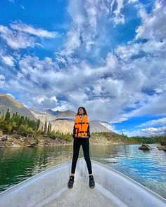 a woman in an orange life jacket standing on the back of a boat with mountains and trees in the background