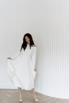 a woman standing in front of a white wall wearing a white dress and high heels