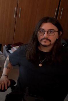 a man with long hair and glasses sitting in front of a computer desk, looking at the camera