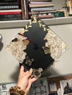 a person holding up a black and gold graduation cap with butterflies on the front, in front of bookshelves