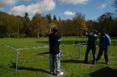 three men are flying a kite in a field with blue tape on the ground and trees behind them