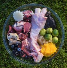 a glass bowl filled with meat and vegetables on top of green grass next to fruit