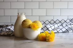 three yellow dandelions in a white bowl next to a bottle of milk on a counter