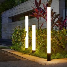 three lights that are sitting in the grass near some bushes and plants on the sidewalk