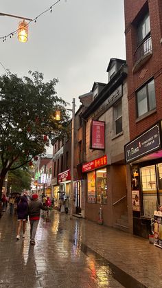 people are walking down the street in front of stores on a rainy day with lights strung overhead