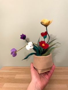 a hand is holding a flower pot with fake flowers in it on a wooden table