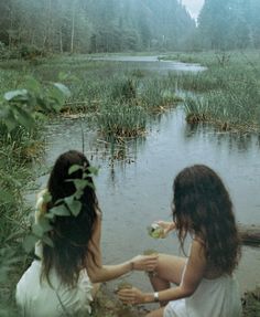 two women sitting on the bank of a river eating food and drinking water from a cup