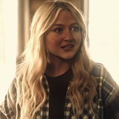 a woman with long blonde hair wearing a black and white checkered shirt standing in front of a window