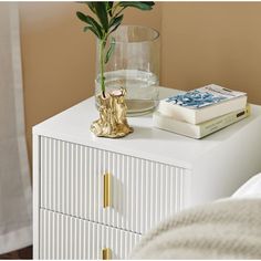 a white nightstand with a glass vase and books on it next to a gold statue