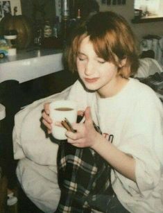 a young person sitting in a kitchen holding a cup of coffee and looking at the camera