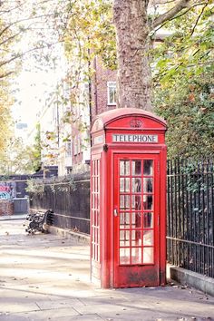 a red telephone booth sitting on the side of a road next to a tree and fence