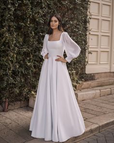 a woman standing in front of a bush wearing a white dress with long sleeves and an open back