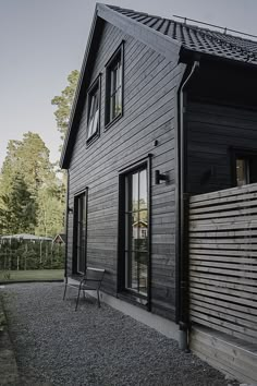 an empty chair sitting outside of a black wooden house in the evening sun on gravel