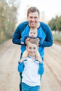 a man holding two small children in his arms and smiling at the camera while standing on a dirt road