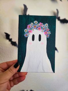 a hand holding up a card with a ghost on it's face and flowers in her hair