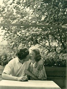 an old photo of two people sitting at a table with one kissing the other's cheek