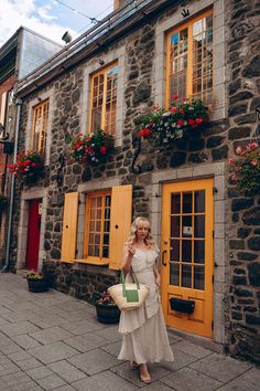 a woman standing in front of a stone building with yellow doors and flowers on the windows