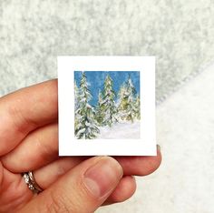 a hand holding a small card with snow covered trees in the background on a white surface