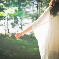 a woman in a white dress blowing bubbles into the air while standing in front of trees