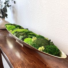 a wooden table topped with a bowl filled with green moss covered plants next to a vase full of flowers