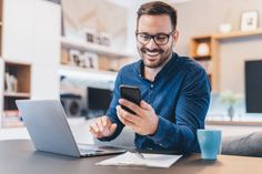 a man sitting at a table looking at his cell phone and smiling while he is on his laptop